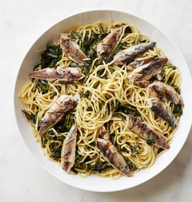 A plate of bucatini pasta topped with grilled sardine fillets, wilted dandelion greens, and frisée, finished with lemon zest and grated Parmesan