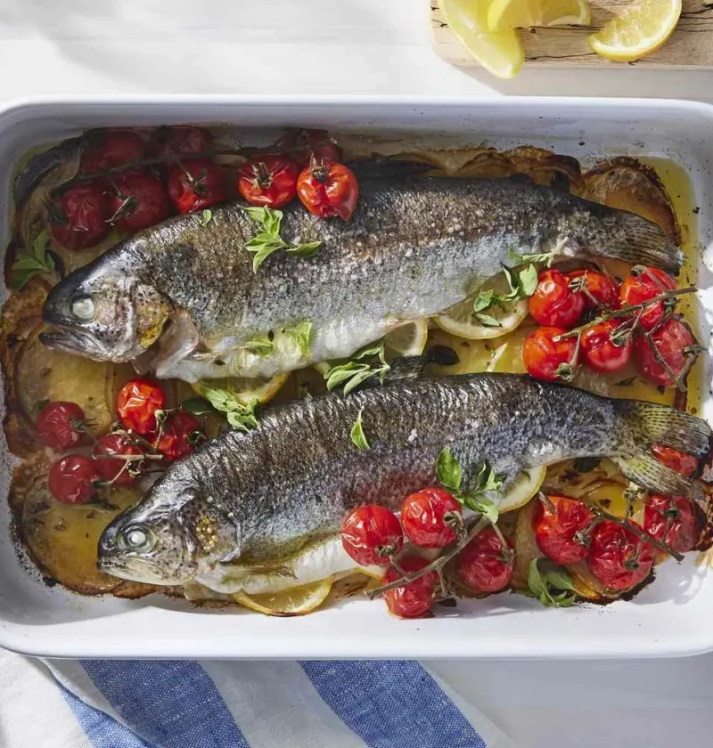 Whole baked trout with crispy skin on a bed of sliced Yukon Gold potatoes and burst cherry tomatoes in a baking dish