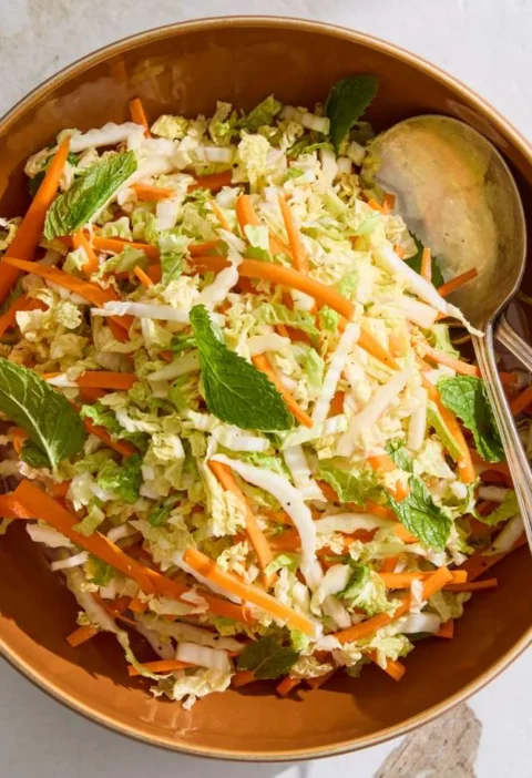 A bowl of Martha Stewart napa cabbage slaw with shredded cabbage, carrot matchsticks, and torn mint leaves in a sesame soy dressing