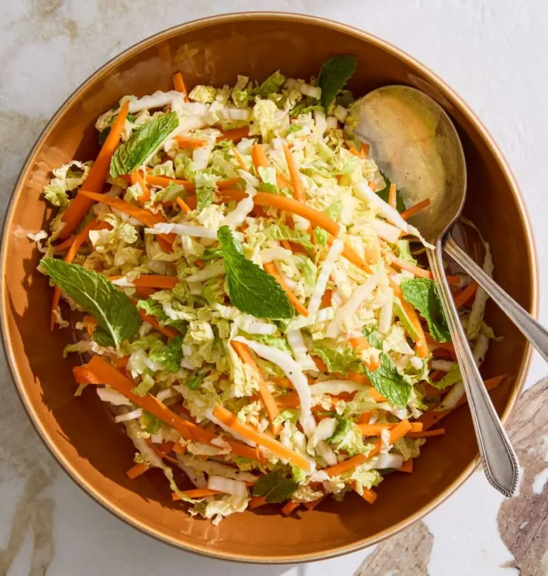 A bowl of Martha Stewart napa cabbage slaw with shredded cabbage, carrot matchsticks, and torn mint leaves in a sesame soy dressing