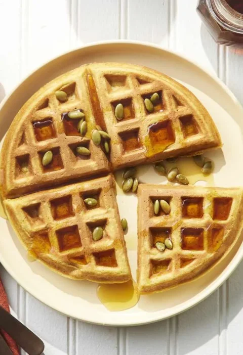 Golden pumpkin waffles stacked on a plate with maple syrup and toasted pumpkin seeds
