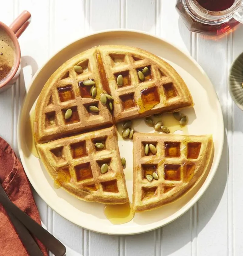 Golden pumpkin waffles stacked on a plate with maple syrup and toasted pumpkin seeds