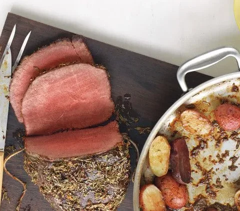 Sliced rosemary-garlic roast beef on a cutting board with roasted potatoes and horseradish sauce