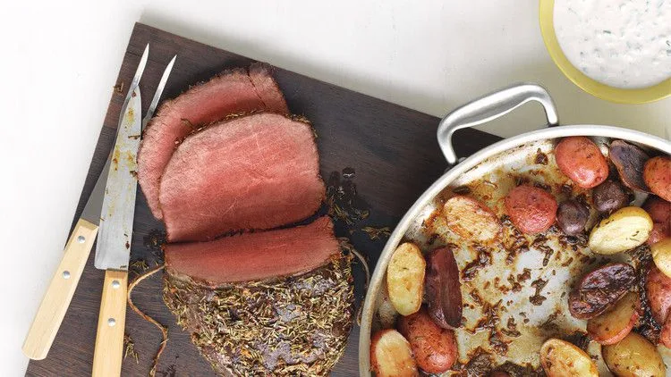 Sliced rosemary-garlic roast beef on a cutting board with roasted potatoes and horseradish sauce