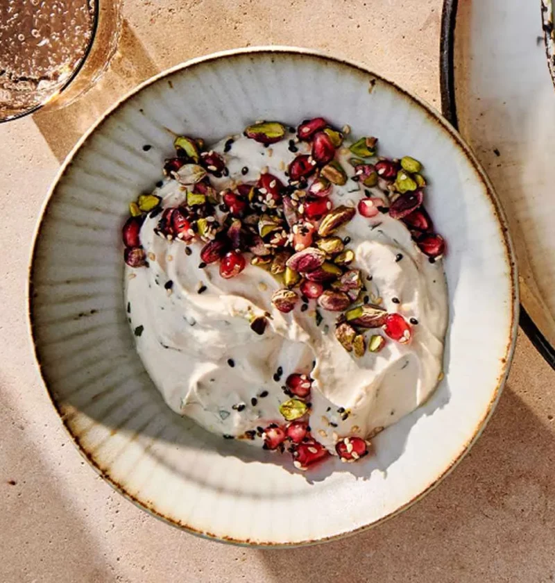 A bowl of creamy tahini-yogurt dip topped with pomegranate arils, toasted pine nuts, and sesame seeds