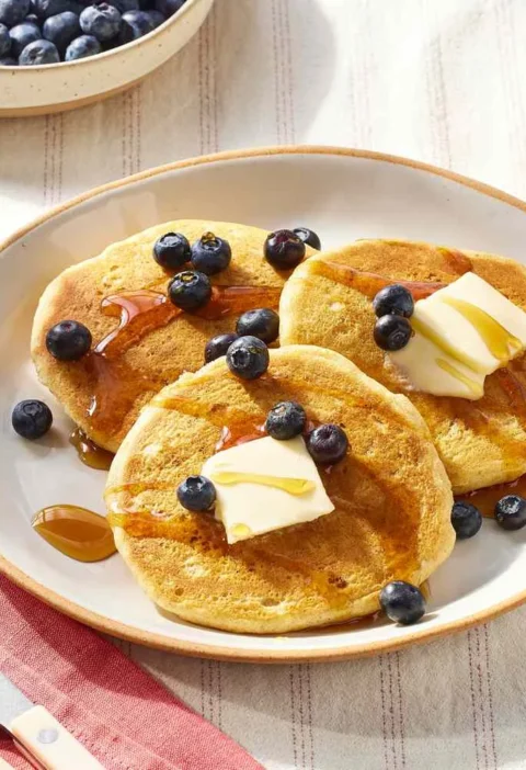 A stack of fluffy golden vegan pancakes drizzled with maple syrup and topped with fresh blueberries
