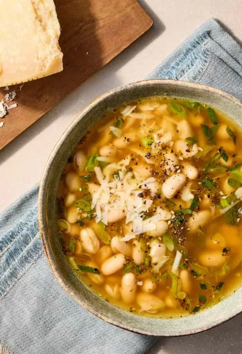 Martha Stewart 15-minute white bean soup in a rustic bowl topped with grated Parmesan, scallions and black pepper next to a wedge of Parmesan