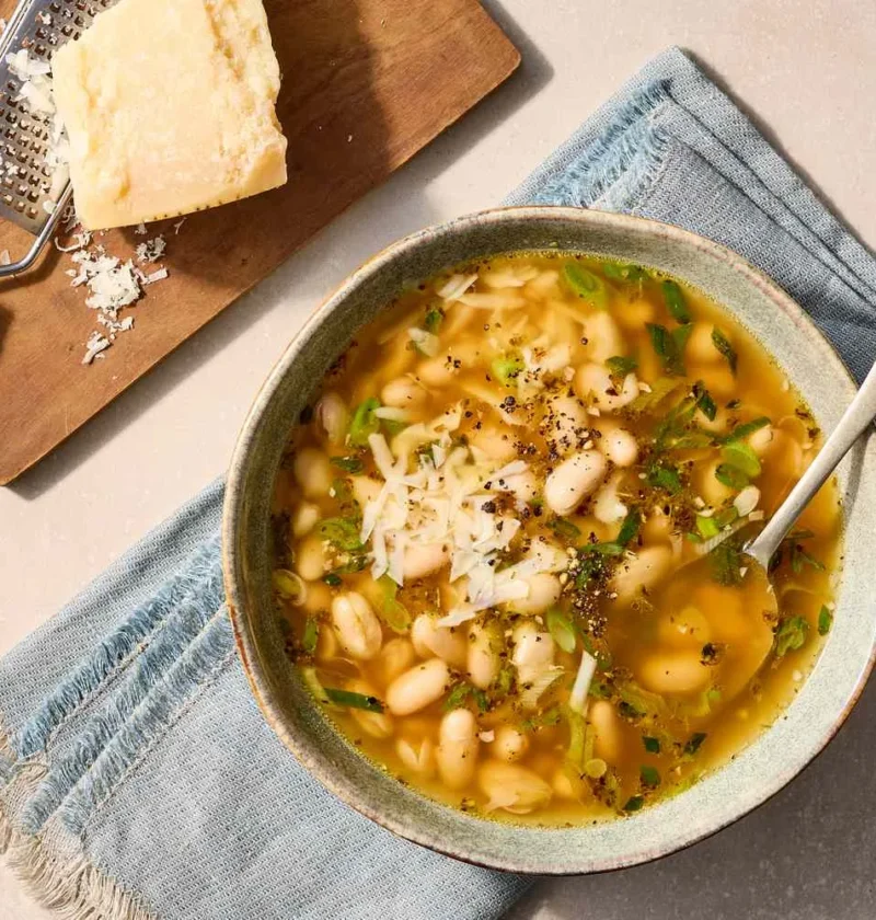 Martha Stewart 15-minute white bean soup in a rustic bowl topped with grated Parmesan, scallions and black pepper next to a wedge of Parmesan