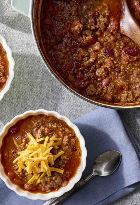 Martha Stewart 30-minute chili topped with shredded cheddar in white bowls next to a Dutch oven of beef and bean chili