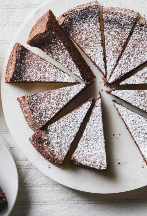 Martha Stewart 4-ingredient flourless chocolate cake cut into slices and dusted with confectioners' sugar next to a bowl of whipped cream