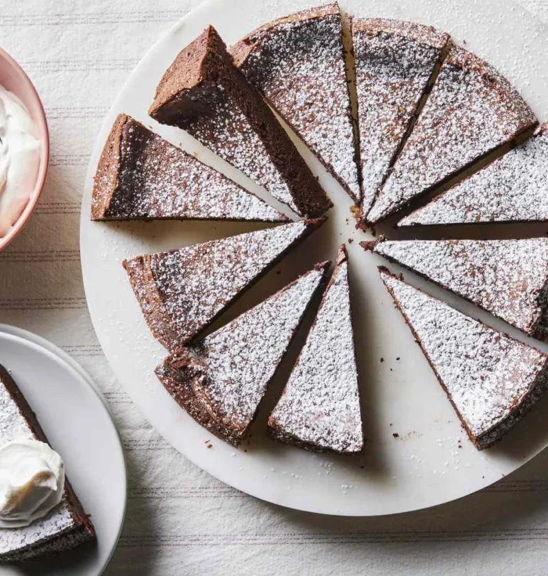 Martha Stewart 4-ingredient flourless chocolate cake cut into slices and dusted with confectioners' sugar next to a bowl of whipped cream