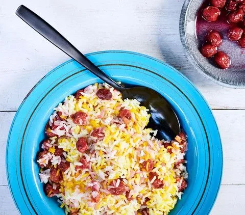Albaloo polo, saffron basmati rice layered with sour cherries, served in a blue bowl with a dark spoon beside a small bowl of cherries