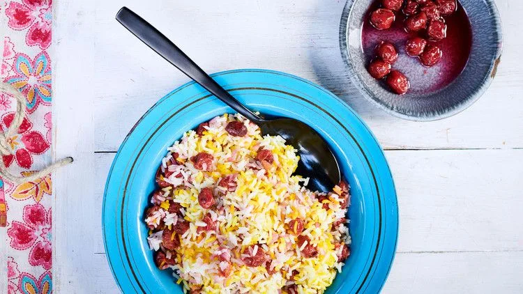 Albaloo polo, saffron basmati rice layered with sour cherries, served in a blue bowl with a dark spoon beside a small bowl of cherries