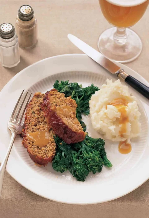 Martha Stewart All-American meatloaf sliced on a white plate with gravy, mashed potatoes and broccoli rabe