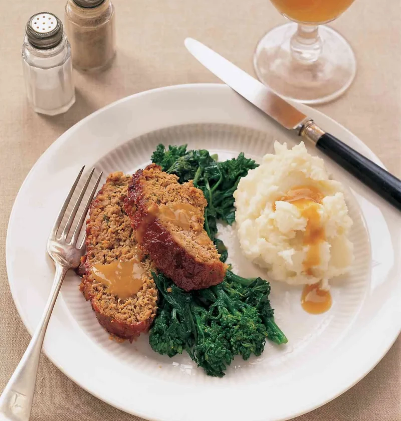 Martha Stewart All-American meatloaf sliced on a white plate with gravy, mashed potatoes and broccoli rabe