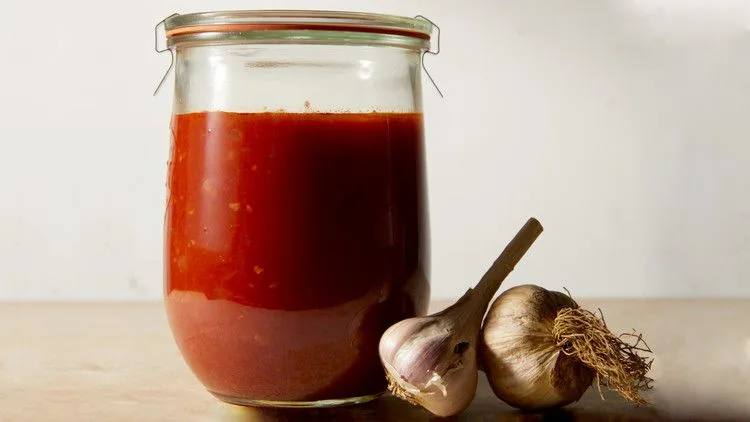 Martha Stewart all-purpose tomato sauce in a glass jar next to two heads of garlic on a wooden surface