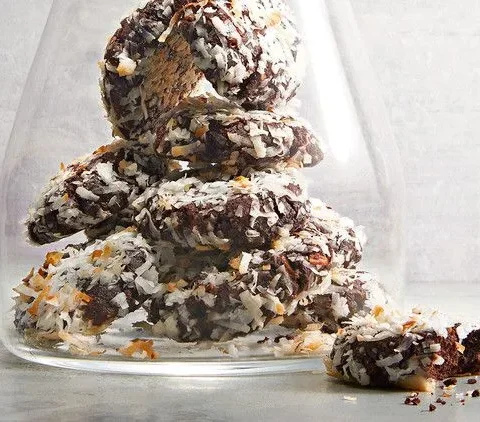 Stack of almond-coconut chewy chocolate cookies coated in shredded coconut inside a glass jar with crumbs nearby