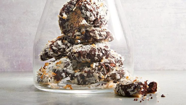 Stack of almond-coconut chewy chocolate cookies coated in shredded coconut inside a glass jar with crumbs nearby