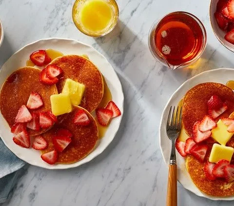 Martha Stewart almond flour pancakes topped with butter and sliced strawberries on white plates with maple syrup