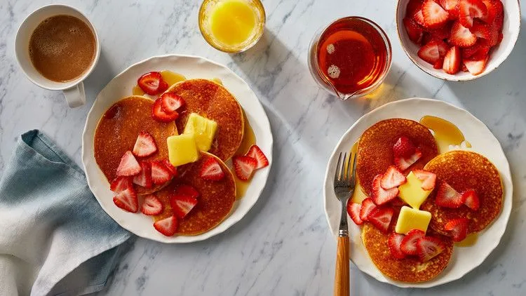 Martha Stewart almond flour pancakes topped with butter and sliced strawberries on white plates with maple syrup