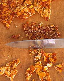 Martha Stewart almond praline broken into shards and finely chopped pieces on a wooden board next to a knife