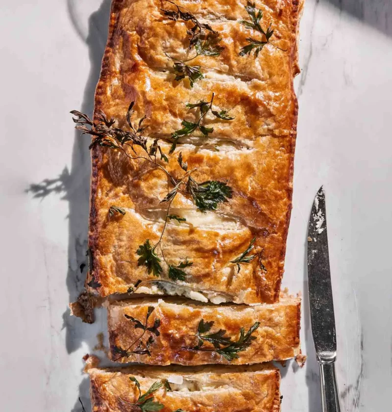 Martha Stewart Alsatian potato pie, a golden puff pastry pie topped with parsley sprigs, sliced on a marble surface