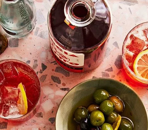Martha Stewart amaro spritz in a glass with ice and a lemon slice beside a bowl of green olives and an orange spritz