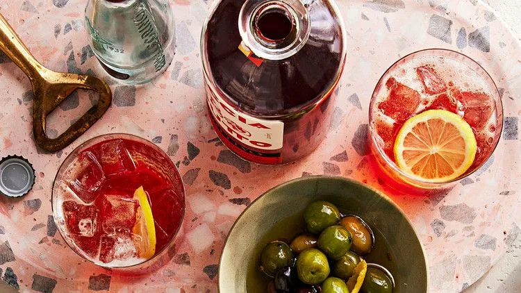 Martha Stewart amaro spritz in a glass with ice and a lemon slice beside a bowl of green olives and an orange spritz