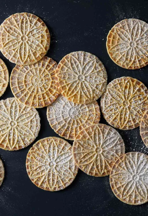 Martha Stewart anise pizzelle cookies with a floral pattern dusted with confectioners' sugar on a dark surface