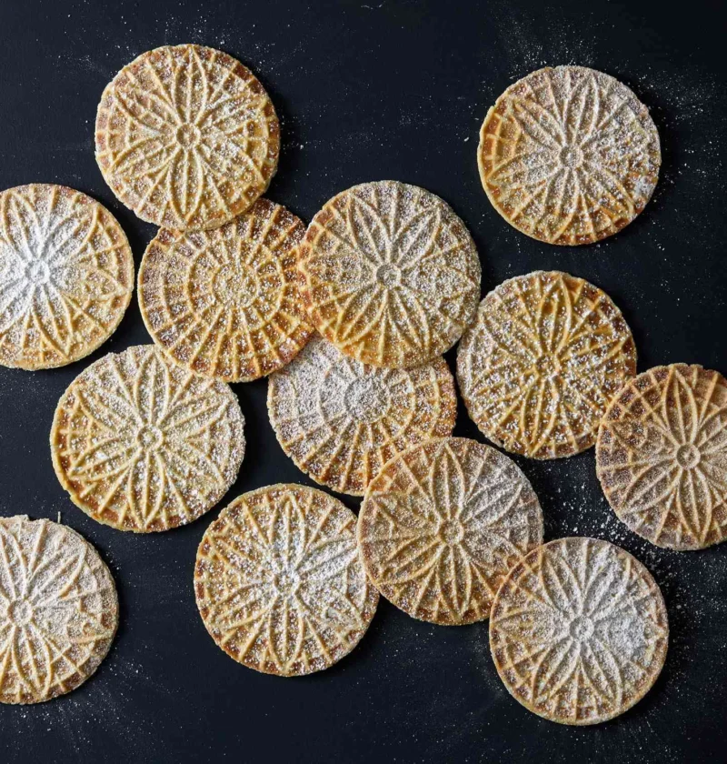 Martha Stewart anise pizzelle cookies with a floral pattern dusted with confectioners' sugar on a dark surface