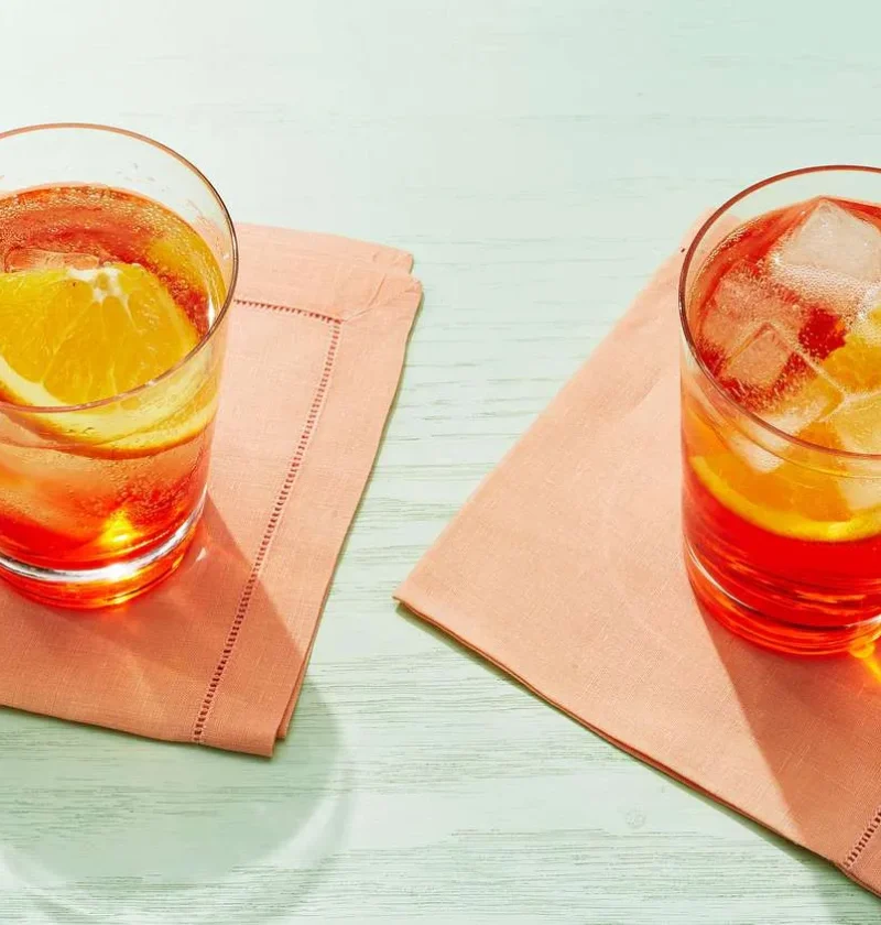 Two Martha Stewart Aperol Spritz drinks with ice and orange slices in glasses on peach napkins