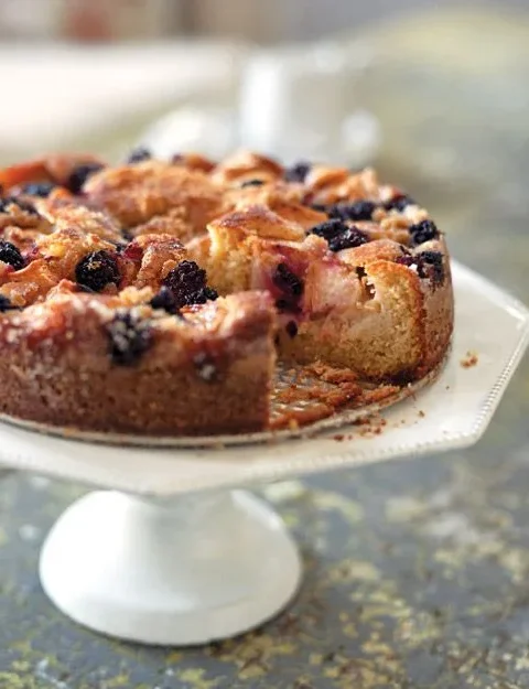Martha Stewart apple-blackberry cake with a slice removed on a white cake stand, topped with baked apple wedges and blackberries