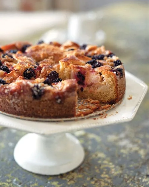 Martha Stewart apple-blackberry cake with a slice removed on a white cake stand, topped with baked apple wedges and blackberries
