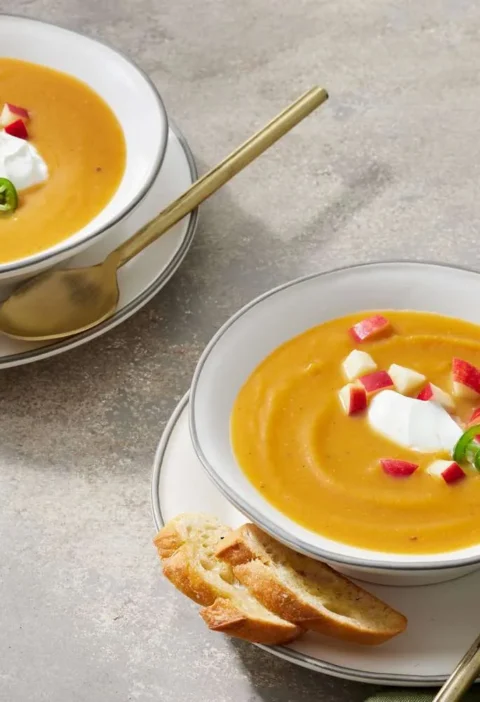 Two bowls of Martha Stewart apple-butternut squash soup topped with diced red apple, jalapeño slices and sour cream, served with toasted bread