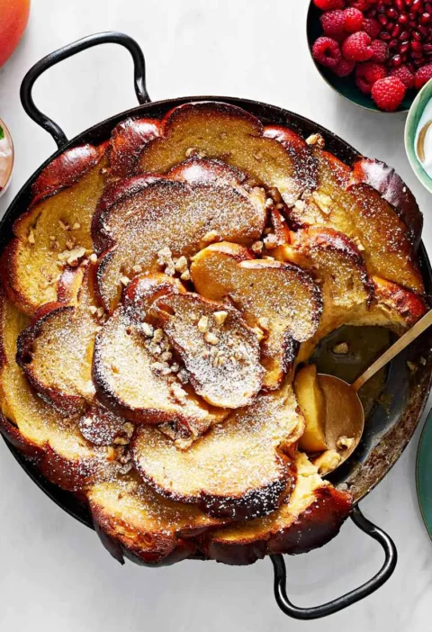 Martha Stewart apple-caramel French toast baked in a black skillet with shingled challah, dusted with confectioners' sugar and chopped pecans