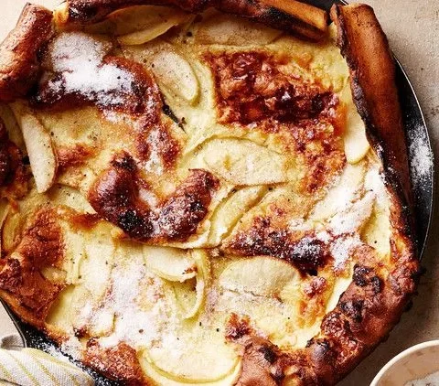 Martha Stewart apple-cardamom brown-butter Dutch baby, puffed and golden in a skillet with apple slices and powdered sugar on top