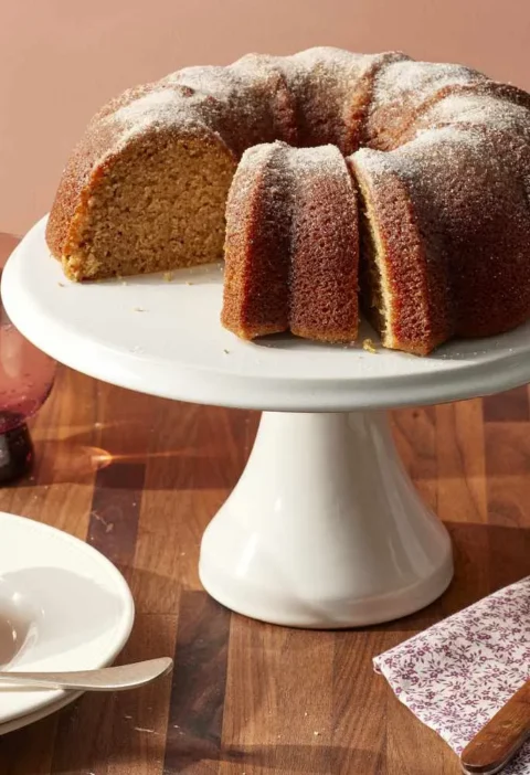 Martha Stewart apple-cider donut cake on a white cake stand dusted with cinnamon sugar, with a sliced piece on a plate