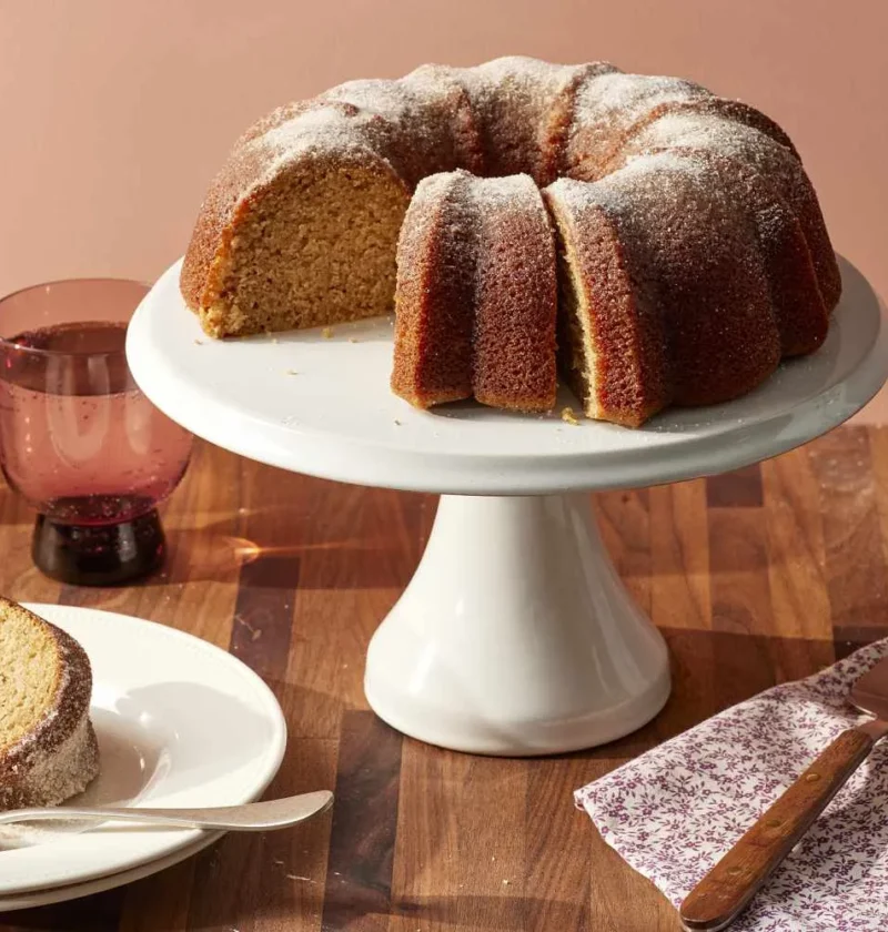 Martha Stewart apple-cider donut cake on a white cake stand dusted with cinnamon sugar, with a sliced piece on a plate