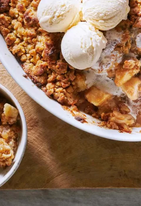Martha Stewart apple crumble in an oval white baking dish topped with scoops of vanilla ice cream beside a bowl of crumble