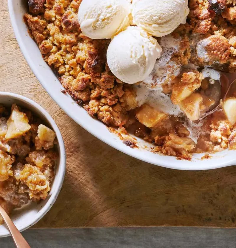 Martha Stewart apple crumble in an oval white baking dish topped with scoops of vanilla ice cream beside a bowl of crumble