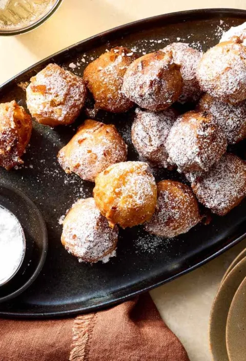Martha Stewart apple fritters dusted with confectioners' sugar on a dark oval platter with a small sifter of powdered sugar