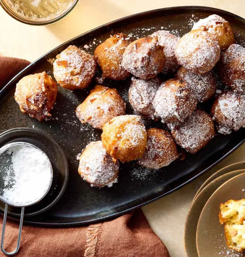 Martha Stewart apple fritters dusted with confectioners' sugar on a dark oval platter with a small sifter of powdered sugar