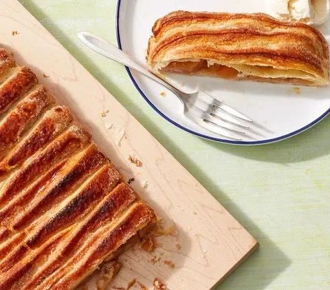 Martha Stewart apple jalousie tart with woven golden puff pastry strips on a wooden board beside a slice and a fork on a white plate