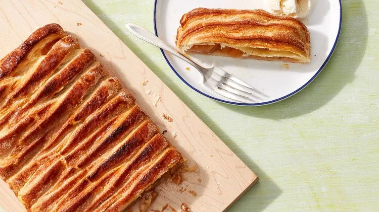 Martha Stewart apple jalousie tart with woven golden puff pastry strips on a wooden board beside a slice and a fork on a white plate