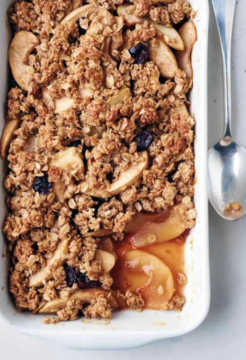 Martha Stewart apple oat crisp with golden oat topping, apple slices and dried cherries in a white baking dish with a spoon