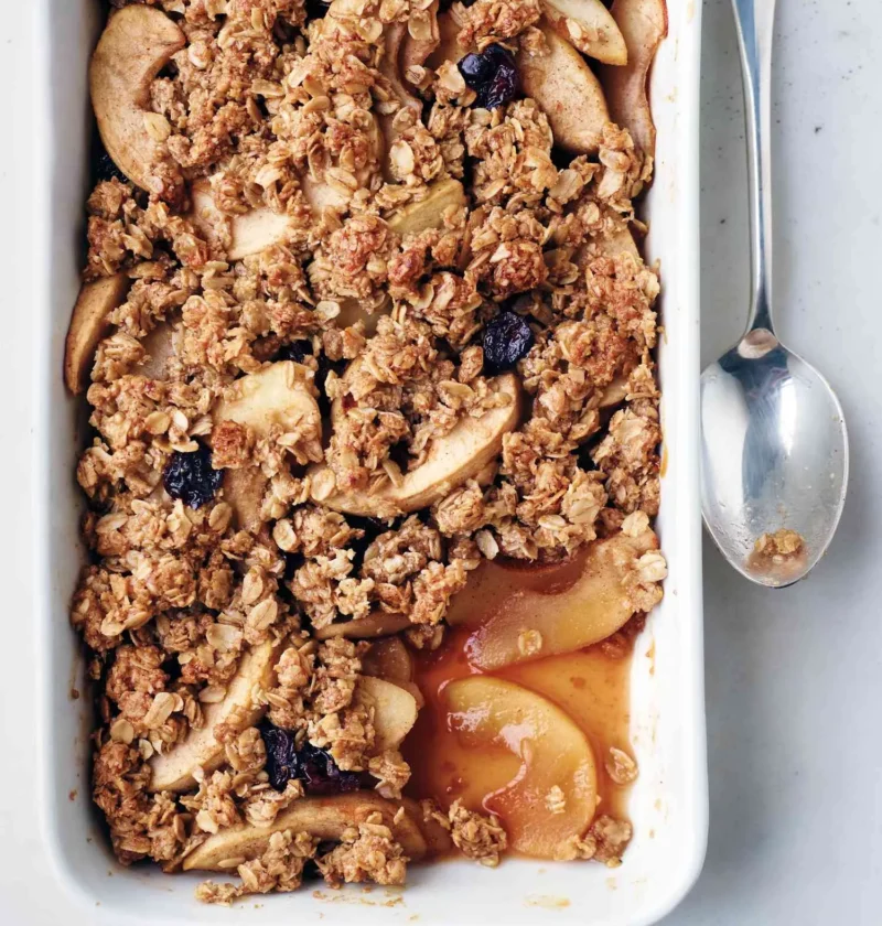Martha Stewart apple oat crisp with golden oat topping, apple slices and dried cherries in a white baking dish with a spoon
