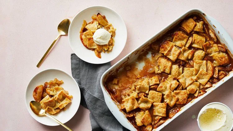 Martha Stewart apple pandowdy in a white baking dish topped with golden pastry squares, with two plates and a scoop of vanilla ice cream beside it