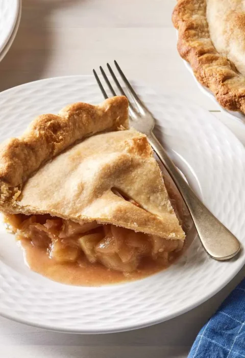 Slice of Martha Stewart apple pie on a white plate with a fork, next to the rest of the golden pie