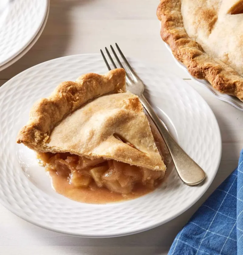 Slice of Martha Stewart apple pie on a white plate with a fork, next to the rest of the golden pie