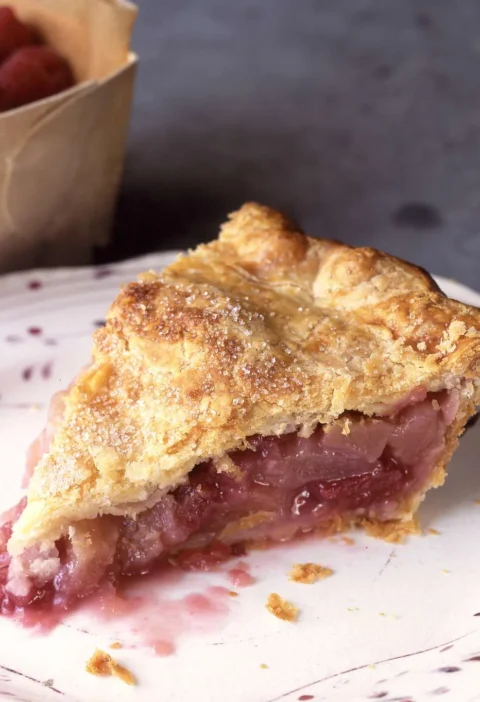 Slice of Martha Stewart apple-raspberry pie with a golden sugar-sprinkled crust and a juicy pink fruit filling on a speckled plate